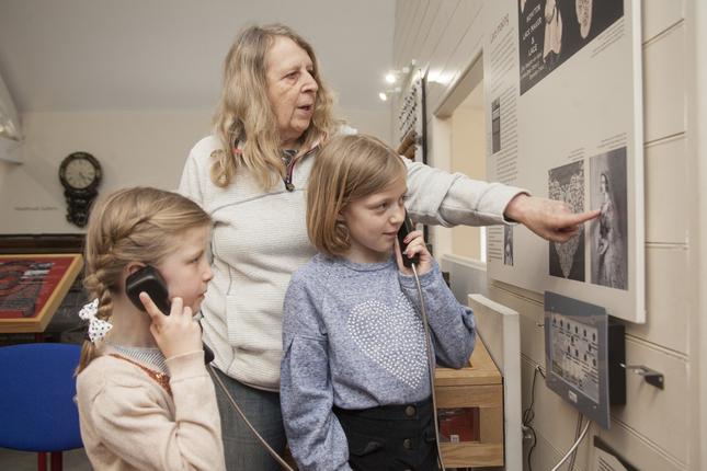 Two girls holding wired telephone handsets to their ears with a woman standing behind them