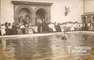 Two men in a swimming pool. One is dragging the other backwards through the water demonstrating an old lifesaving technique to onlookers.