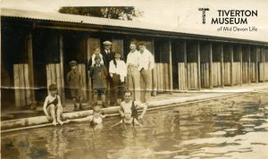 A group of people in a swimming pool looking at the camera with several more standing behind them on the poolside in front of a row of changing cubicles