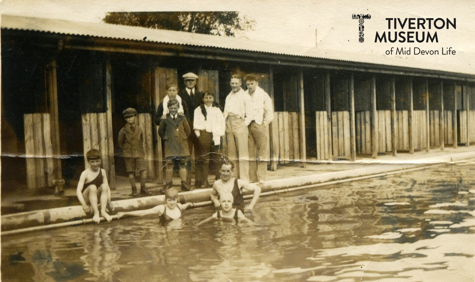 A group of people in a swimming pool looking at the camera with several more standing behind them on the poolside in front of a row of changing cubicles