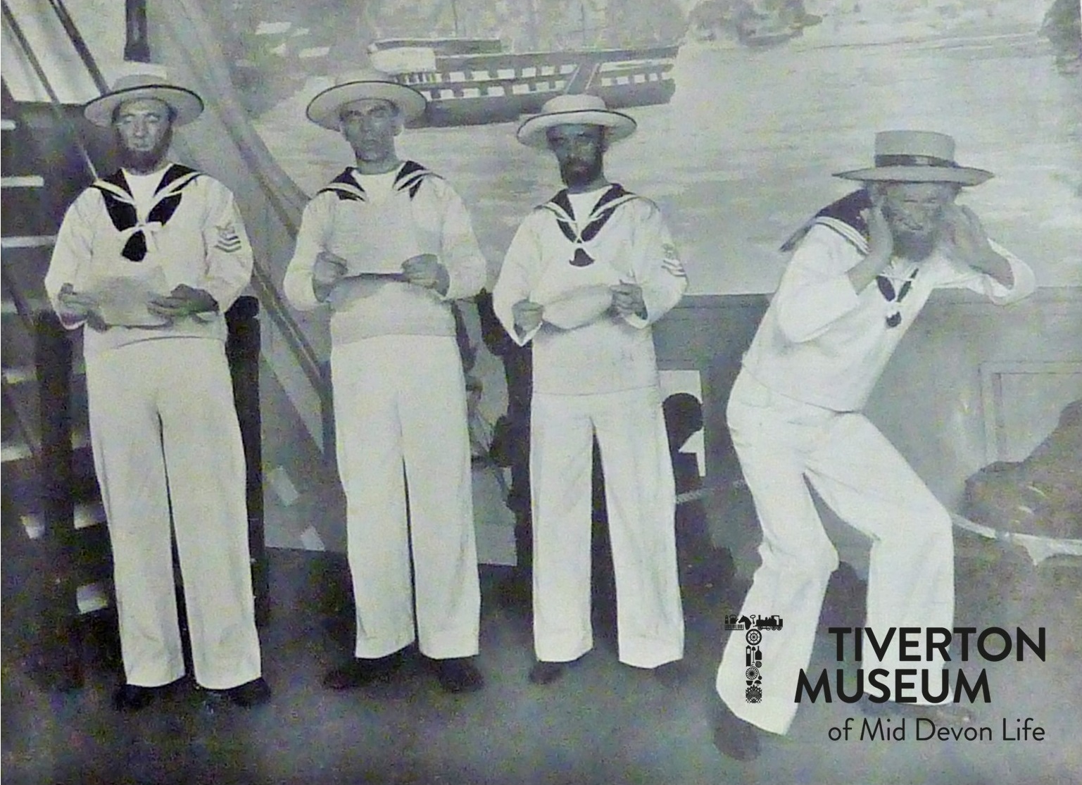 A black and white photo with four men dressed in white sailor uniforms and hats in front of a theatre set.