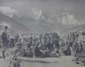 A grainy black and white photo that appears to be from a newspaper. The peaks of Everest loom large in the distant background of a l;arge group of Sherpa porters and guides sitting and standing in the foreground. Some are looking at the camera, others are engaged in what they're doing. 