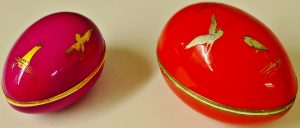 Two egg shaped boxes. One is red or orange, the other is pink and they both have a gold trim at the join which runs vertically through the middle of the egg. They are both painted with simple gold bird outlines. 