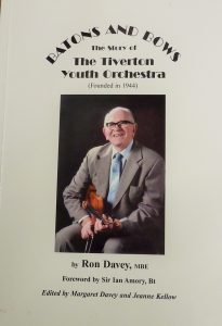 The front cover of a book called Batons and Bows, the story of the Tiverton Youth Orchestra, featuring a photo of a seated man with a big smile, holding a violin. 