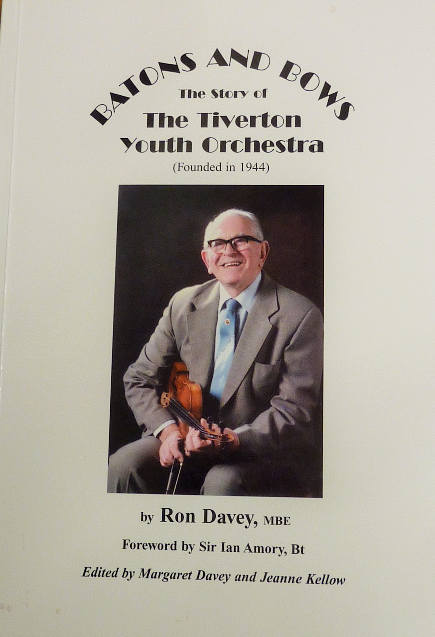 The front cover of a book called Batons and Bows, the story of the Tiverton Youth Orchestra, featuring a photo of a seated man with a big smile, holding a violin.