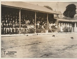 An open air swimming pool with several children swimming towards the camera. There are stands in the background with lots of children watching.