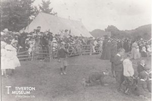 Large crowds of people in a country show ground with a marquee in the background. The people are dressed in old fashioned clothing, dresses for the women and girls, short trousers for the boys and suits for the men. most are wearing hats. 