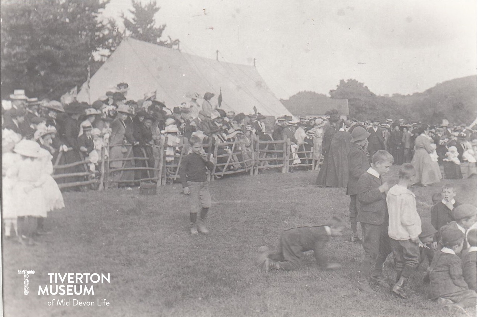 Large crowds of people in a country show ground with a marquee in the background. The people are dressed in old fashioned clothing, dresses for the women and girls, short trousers for the boys and suits for the men. most are wearing hats.