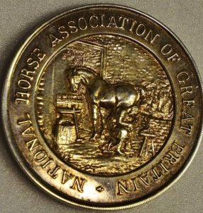 The front of a medal which still looks quite shiny. It has an embossed engraving of a farrier in a blackmisths shop shoeing a horse. In a circle around the outside of the medal it reads 'National Horse Association of Great Britain'