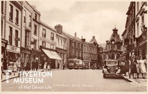 A postcard showing Tiverton's Fore Street in the 1950s. A street scenes with buildings, several old fashioned cars on the street and people walking on the pavements. The women seem to have summer dresses on. 