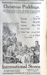 Newspaper advert for Christmas Pudding ingredients at International Stores. There is an illustration of a family around a Christmas dinner table about to be served a Christmas pudding by a servant. 