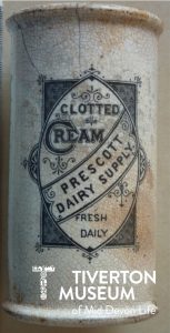 A close up of a ceramic Clotted cream container. It's white in colour, with a cracked glaze, and looks like it is tall and thin. An embellished design in black ink on the front of the container reads 'Clotted Cream - Prescott Dairy Supply - Fresh Daily'.