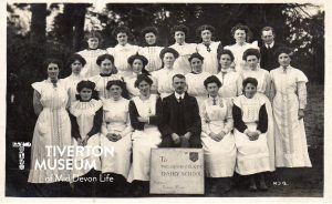 A group of young white women with Edwardian hair styles wearing long white pinafores sitting and standing in rows for the photo. Most of the women are smiling. There is a very serious looking man in the centre front row wearing a dark suit. Resting against his knees there is a board reading 'To The Davon Dairy School. Admission Visitor 3d each'. There is another man in a dark suit, wearing glasses at the back row on the right hand side. He seems to have a little smile on his face. 