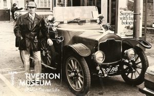 A man standing next to a classic car. The photo is dated 1980 but the car is much older, with big wheels, open top, horn on the side, large lamp headlights. The man is wearing a leather look jacket with belt with shirt and tie, and a deerstalker hat. There's child sitting in the car. In the background there is a bus shelter with an advert that appears to read 'surprise him with faggots in bolognese'. 