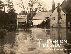 Flood waters in front of several buildings and gates. There is a sign in front of the gate that reads 'Tiverton Motor Co'. The flood looks quite deep. 